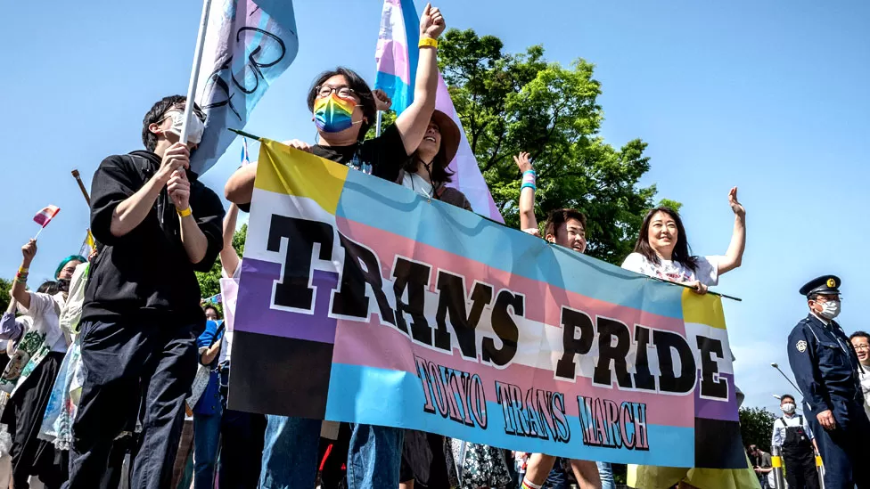 A group of young Japanese hold a large hanging flag reading Trans Pride. The line below that reads Tokyo Trans March